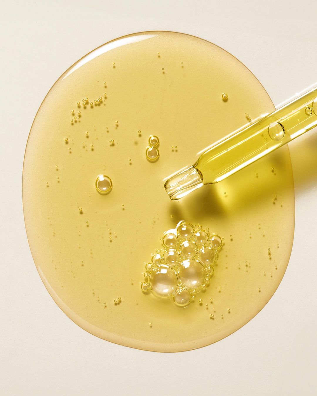 Overhead shot of cosmetic serum with bubbles and a pipette on an off-white background, showcasing texture and product consistency.