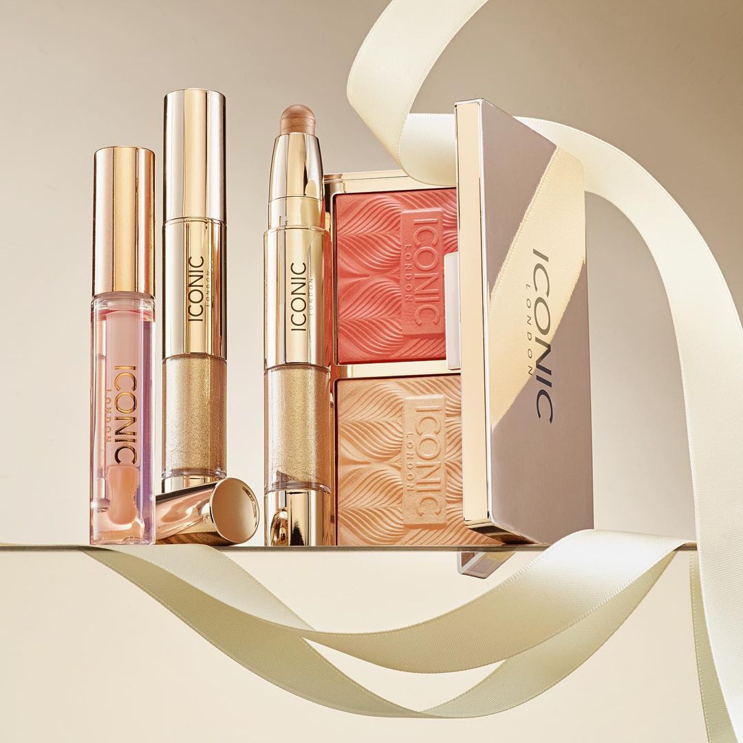 Still life of Iconic London makeup products featuring lipstick, blush palettes, and highlighter on a reflective surface with cream ribbon.