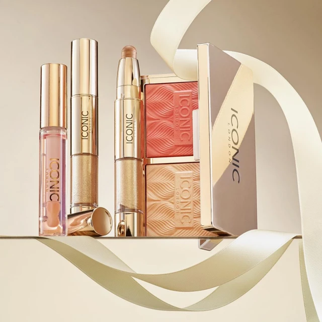 Still life of Iconic London makeup products featuring lipstick, blush palettes, and highlighter on a reflective surface with cream ribbon.