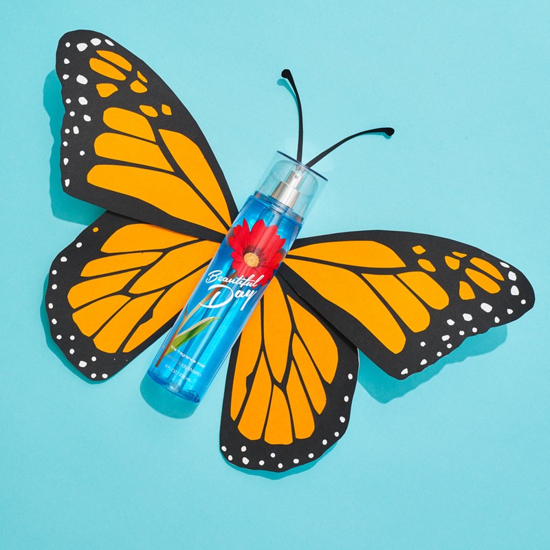 A clear blue bottle labeled 'Beautiful Day' sits against a bright blue backdrop with a decorative butterfly cut-out.