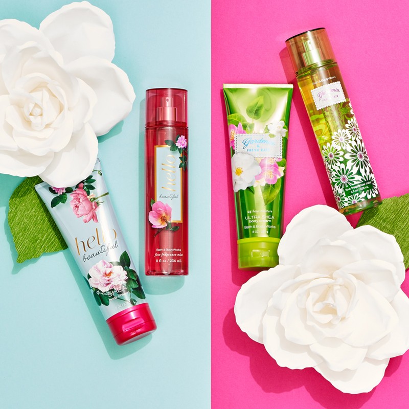 Three Bath & Body Works products are arranged on a split pink and blue background, accented by white flowers and green leaves.