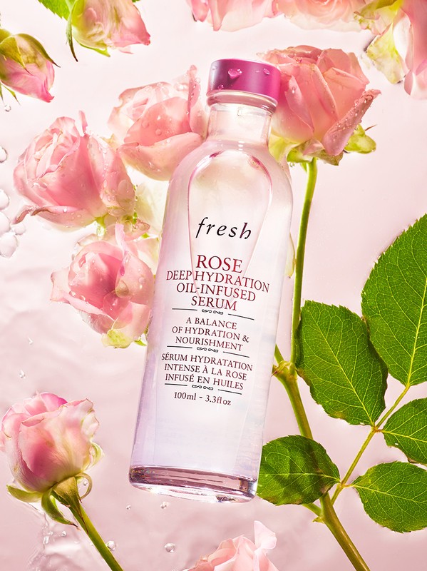 Clear glass bottle of Fresh Rose Deep Hydration Oil-Infused Serum surrounded by pink roses, stems, and leaves on a pink background.