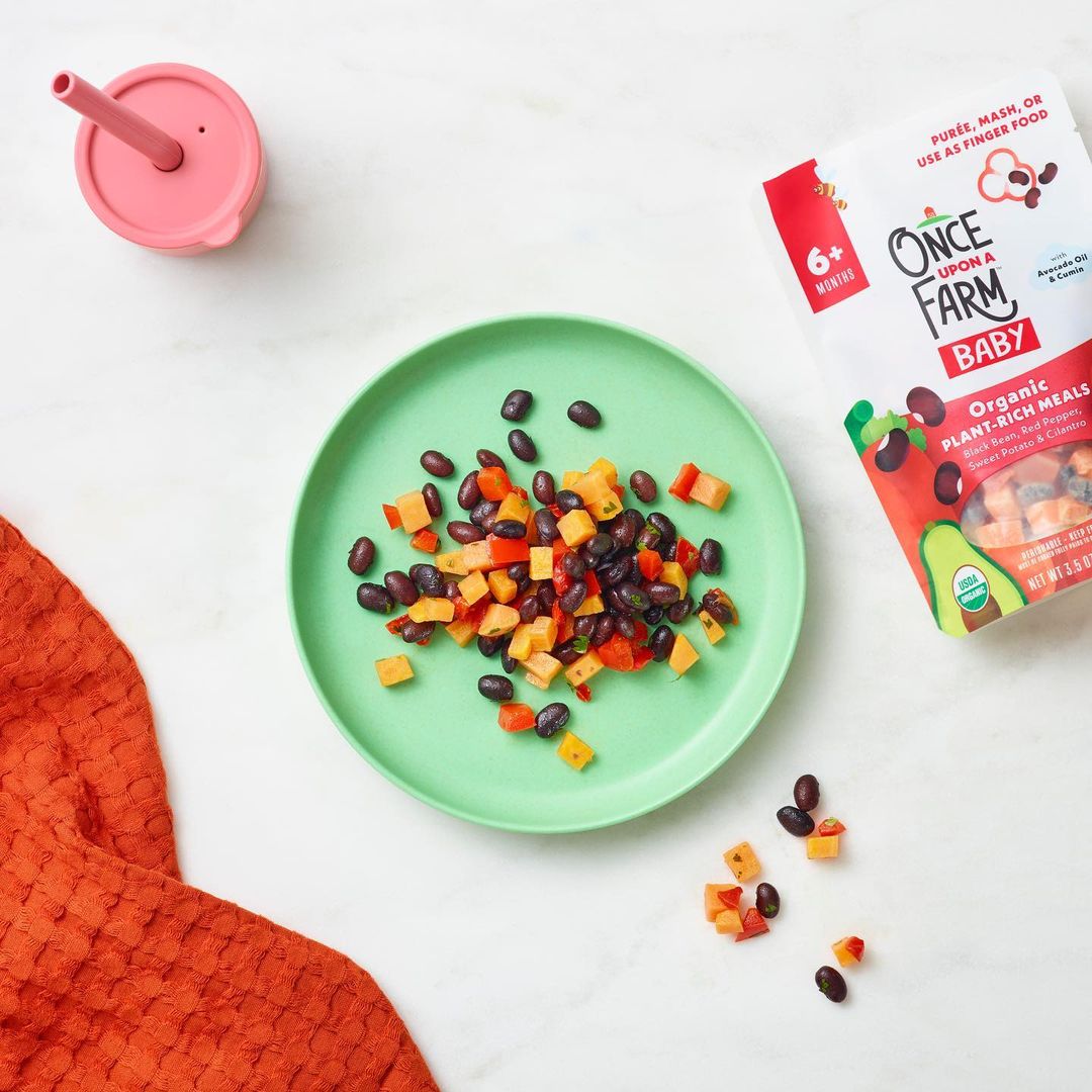 Overhead shot of Once Upon a Farm baby food meal with black beans and sweet potatoes on a plate, featuring a sippy cup and orange towel.