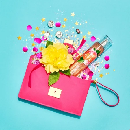 Flat lay photo of a bright pink purse with a yellow flower, bottle of body mist, and confetti on a blue background.