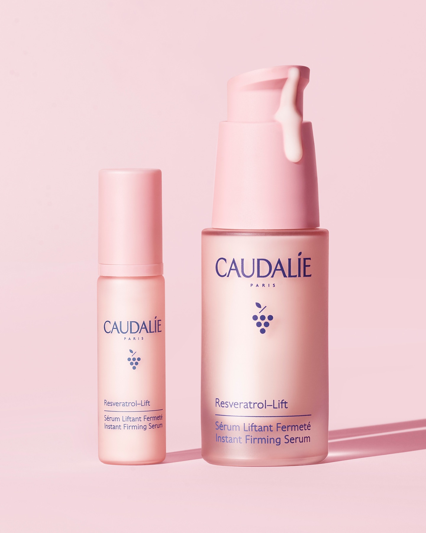Two pink Caudalie Resveratrol Lift skincare product bottles with serum dripping from the pump, on a light pink background in a studio.