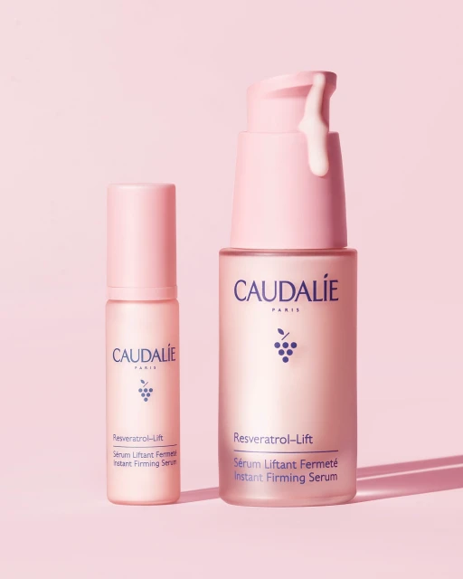 Two pink Caudalie Resveratrol Lift skincare product bottles with serum dripping from the pump, on a light pink background in a studio.