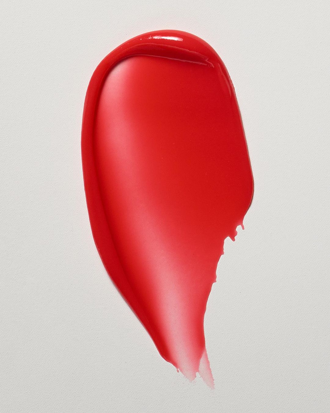 Close-up studio shot of a textured smear of shiny, bright red cosmetic product against a plain white backdrop, beauty advertisement.