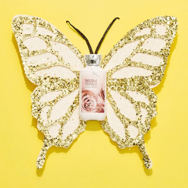 Bath & Body Works Warm Vanilla Sugar lotion bottle on a gold glitter butterfly against a bright yellow background in a studio setting.