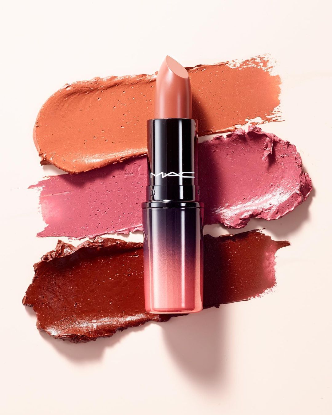 Close-up studio shot of a MAC lipstick tube with a nude lipstick bullet, set against smears of different lipstick shades.
