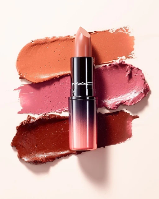 Close-up studio shot of a MAC lipstick tube with a nude lipstick bullet, set against smears of different lipstick shades.