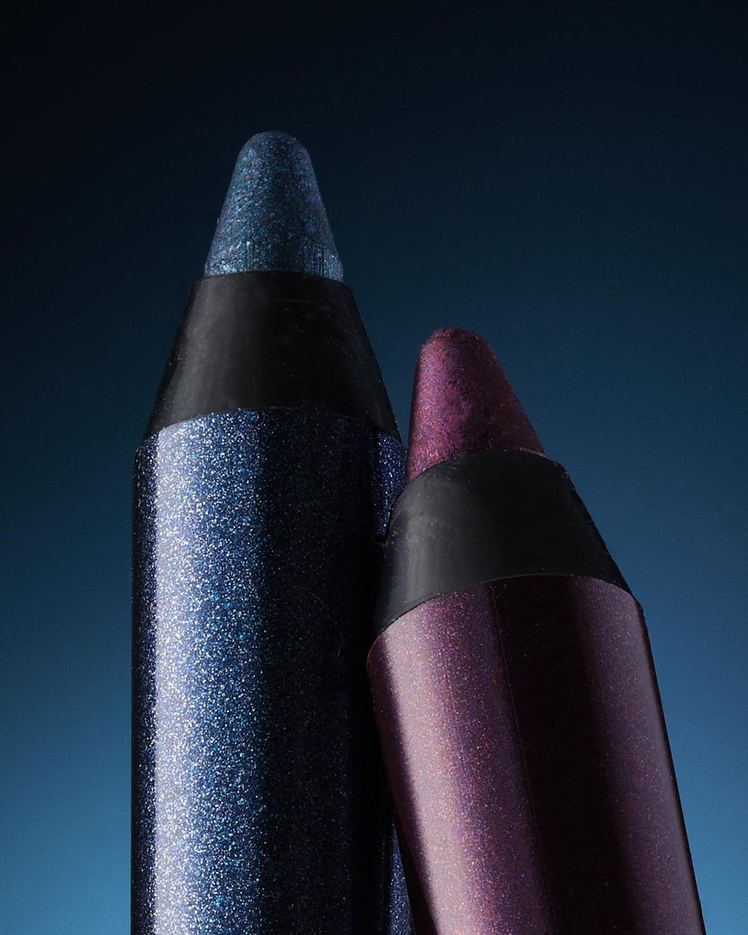 Close-up of two glittery eye makeup pencils, one blue and one maroon, positioned against a gradient blue background in studio lighting.
