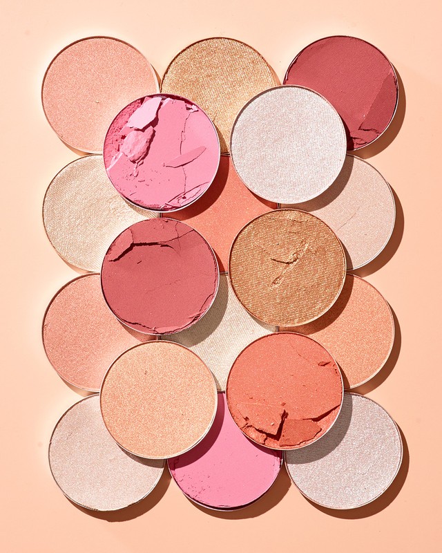 Close-up flat lay of various colors of round blush and highlighter makeup pans arranged on a peach color background.
