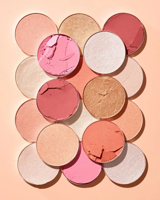 Close-up flat lay of various colors of round blush and highlighter makeup pans arranged on a peach color background.