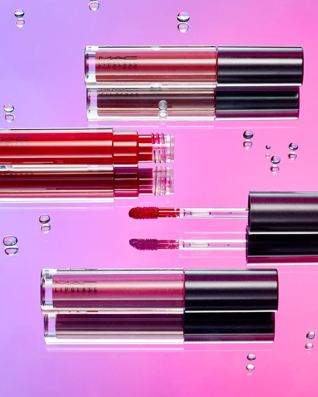 Several tubes of MAC Lipglass in various shades are displayed on a reflective surface with water droplets against a pink and purple gradient.