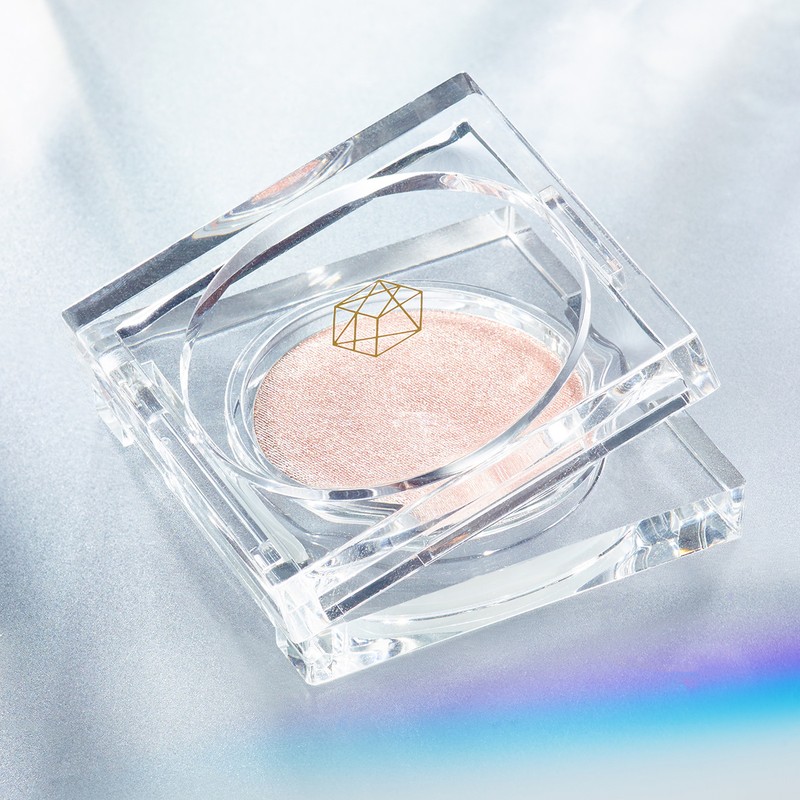 High angle close-up of a reflective, clear acrylic container with a round pressed powder inside on a reflective surface with a rainbow glare.