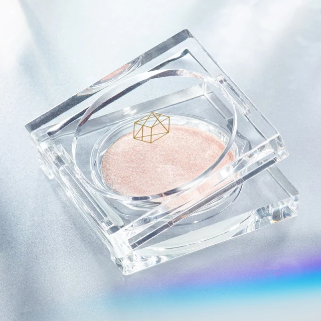 High angle close-up of a reflective, clear acrylic container with a round pressed powder inside on a reflective surface with a rainbow glare.