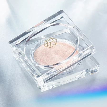 High angle close-up of a reflective, clear acrylic container with a round pressed powder inside on a reflective surface with a rainbow glare.