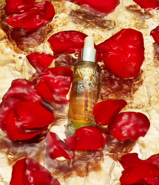 Caudalie Premier Cru serum surrounded by vibrant red rose petals on a reflective, textured gold surface, showcasing a beauty product.