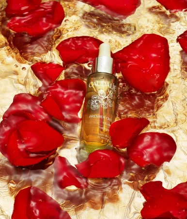 Caudalie Premier Cru serum surrounded by vibrant red rose petals on a reflective, textured gold surface, showcasing a beauty product.