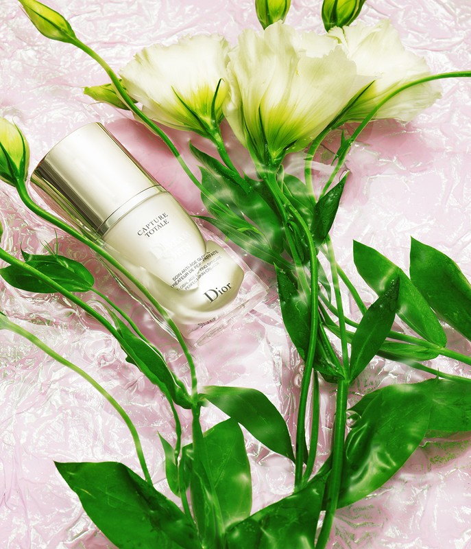 Still life featuring a Dior skincare product amid white flowers and green foliage, set against a textured pink surface, providing a beauty aesthetic.