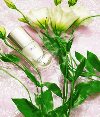 Still life featuring a Dior skincare product amid white flowers and green foliage, set against a textured pink surface, providing a beauty aesthetic.