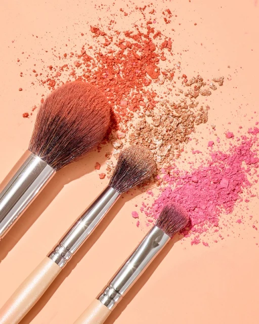 Makeup brushes with silver handles are surrounded by colorful powder on a peach background in a beauty or fashion product shot.