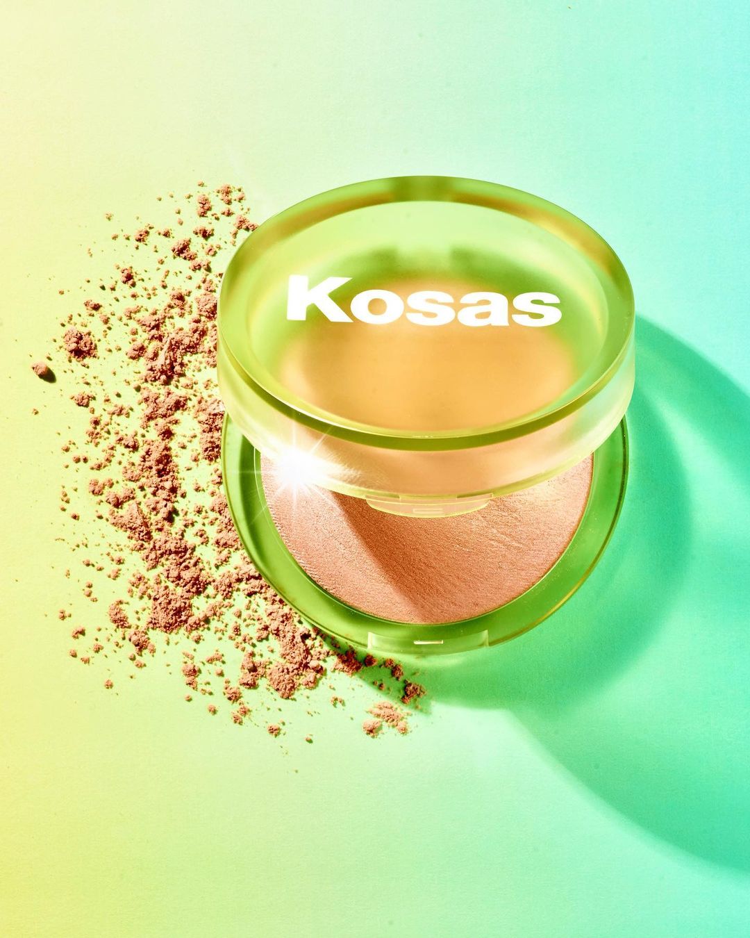 Cosmetic powder foundation by Kosas with loose powder on a bright gradient background in shades of green, yellow, and blue.