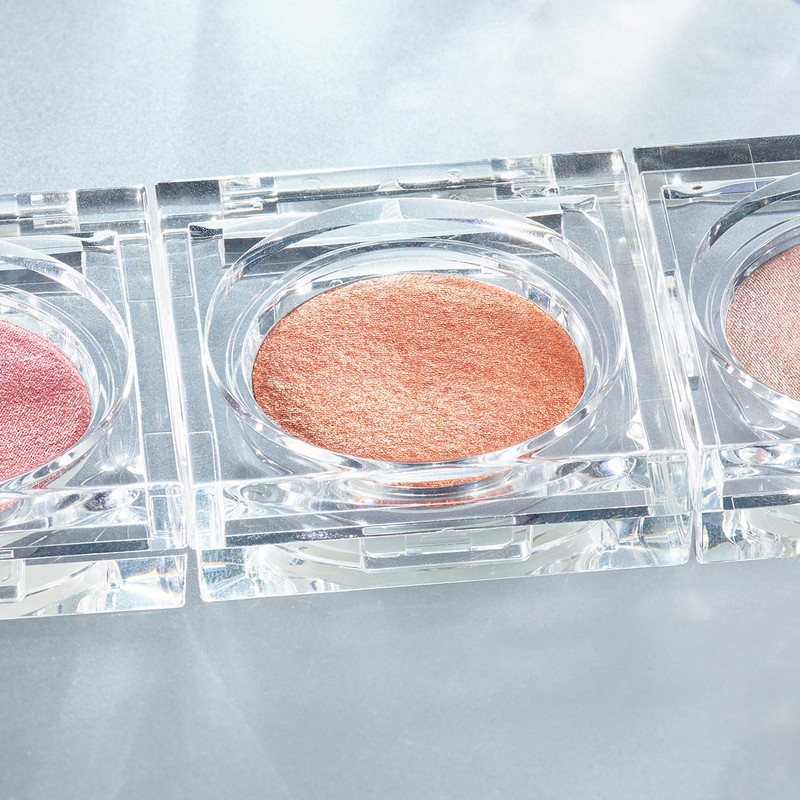 Close-up of three makeup compacts with clear cases and various shades of shimmery powder, set on a surface with diffused lighting.