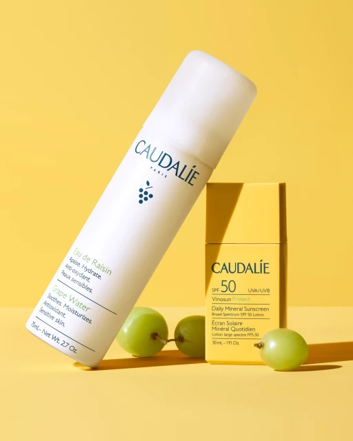Two Caudalie beauty products, a white spray bottle and yellow sunscreen, are displayed with green grapes against a yellow background.