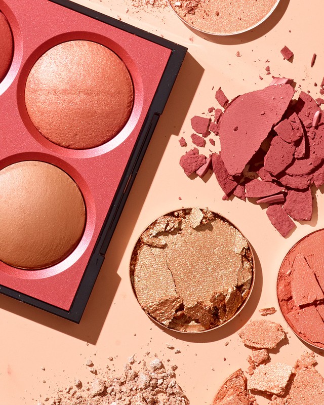 Overhead shot of a makeup palette with bronze and pink blush next to loose, broken powder on a peach-colored surface, beauty product.