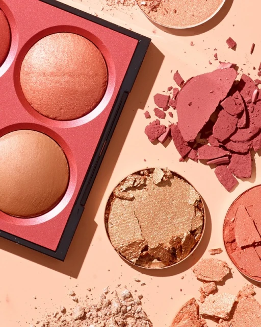 Overhead shot of a makeup palette with bronze and pink blush next to loose, broken powder on a peach-colored surface, beauty product.