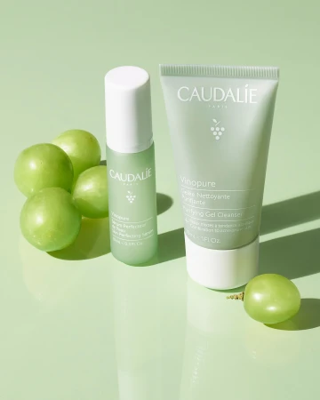 Studio shot of Caudalie Vinopure skincare products including a gel cleanser, serum, and green grapes, on a light green surface.
