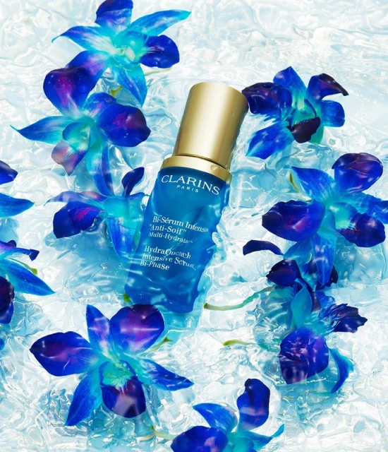 Blue skincare serum bottle with gold cap surrounded by blue orchid flowers against a light blue, textured background in a close-up product shot.