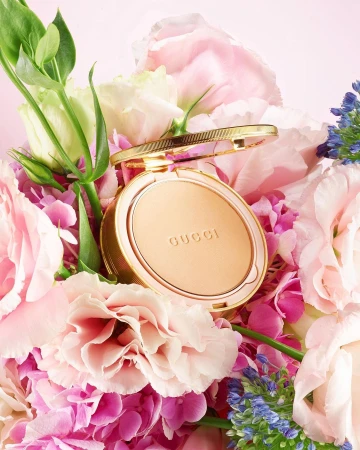 Gucci powder compact on a bed of light pink, blue, and white flowers. The compact is gold with the brand name printed on the powder.