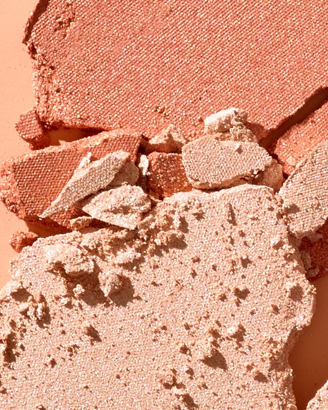 Close-up shot of broken pieces of shimmering peach and tan coloured powder makeup, with a light-peach colored background.
