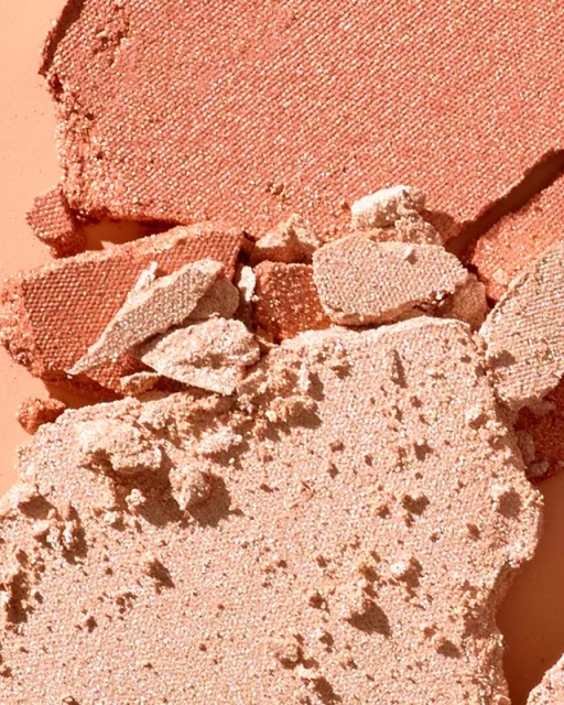 Close-up shot of broken pieces of shimmering peach and tan coloured powder makeup, with a light-peach colored background.