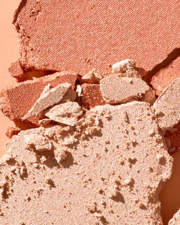 Close-up shot of broken pieces of shimmering peach and tan coloured powder makeup, with a light-peach colored background.
