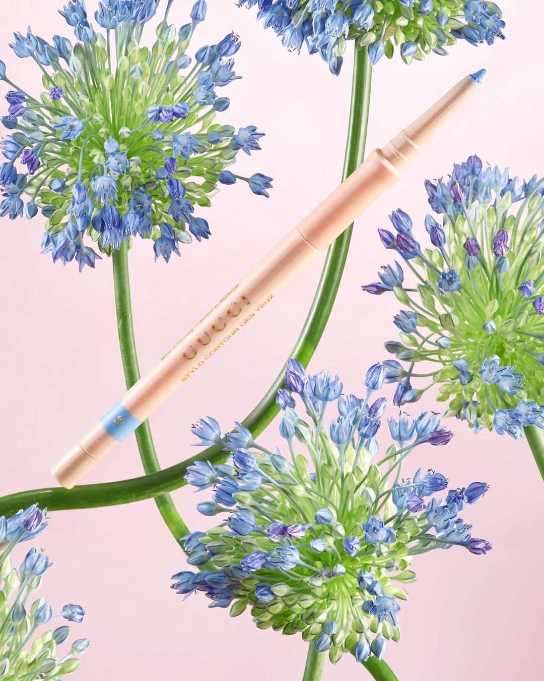 Gucci Stylo Contour des Yeux eyeliner pencil showcased with flowering blue allium blooms against a soft pink backdrop in studio shot.