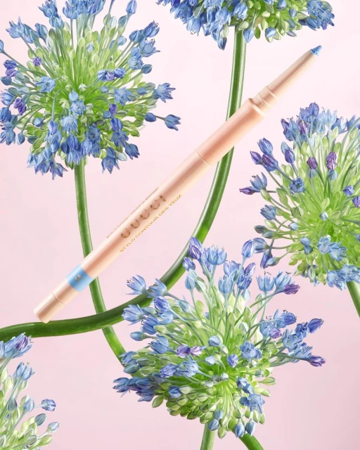 Gucci Stylo Contour des Yeux eyeliner pencil showcased with flowering blue allium blooms against a soft pink backdrop in studio shot.