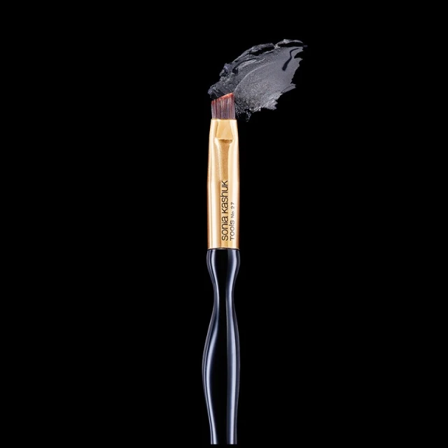 Makeup brush with black handle and gold tone ferrule next to swatch of gray makeup against a solid black background in studio lighting.