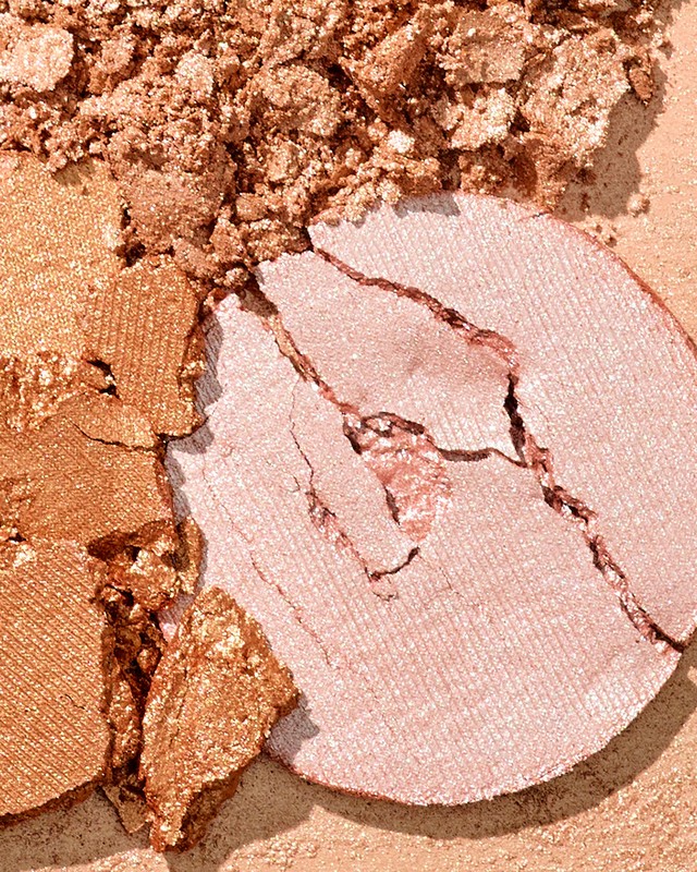 Close-up of a shattered, light-pink makeup powder compact, with an adjacent section of crushed bronze cosmetic powder.