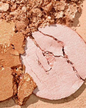 Close-up of a shattered, light-pink makeup powder compact, with an adjacent section of crushed bronze cosmetic powder.