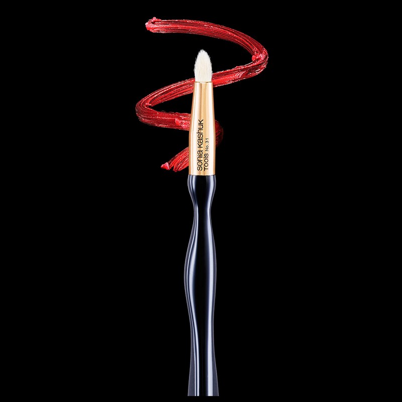 Makeup brush with black handle and synthetic white fibers set against a dark background with smear of red cosmetic product.