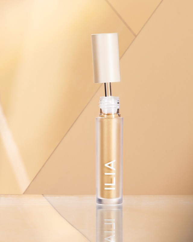 Close-up of an open Ilia liquid eyeshadow bottle with a wand applicator against a beige geometric background, creating an elegant beauty shot.