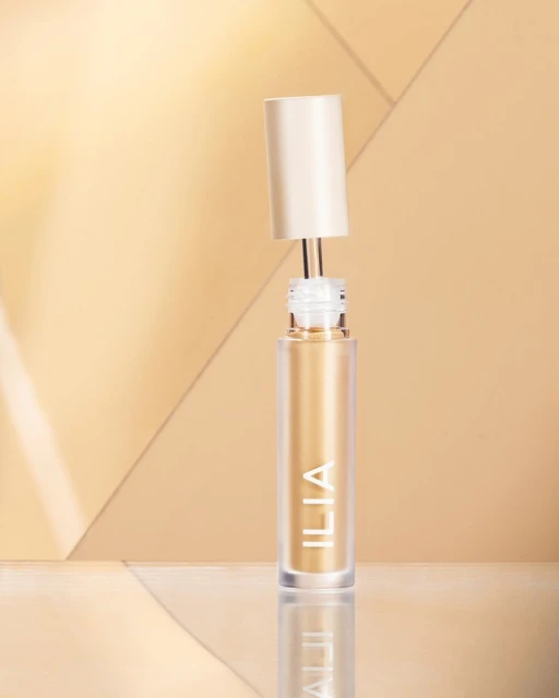 Close-up of an open Ilia liquid eyeshadow bottle with a wand applicator against a beige geometric background, creating an elegant beauty shot.