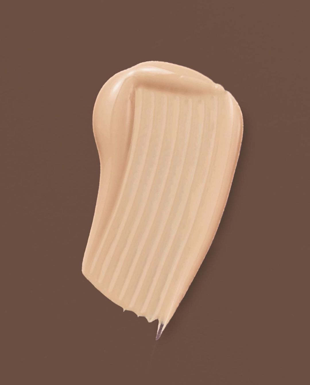 Close-up of light tan makeup foundation smear on brown background, showing its creamy texture and smooth appearance.