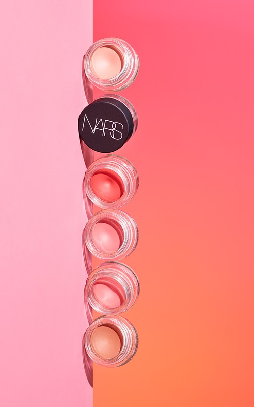 Stack of NARS brand cream makeup in clear jars with pink, peach, and orange colors, set against pink and orange background.