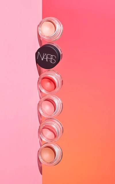 Stack of NARS brand cream makeup in clear jars with pink, peach, and orange colors, set against pink and orange background.