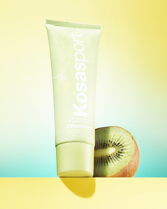 Product shot of Kosasport Chemistry AHA Serum Deodorant in a green tube alongside a halved kiwi, set against a yellow and teal background.