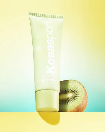 Product shot of Kosasport Chemistry AHA Serum Deodorant in a green tube alongside a halved kiwi, set against a yellow and teal background.